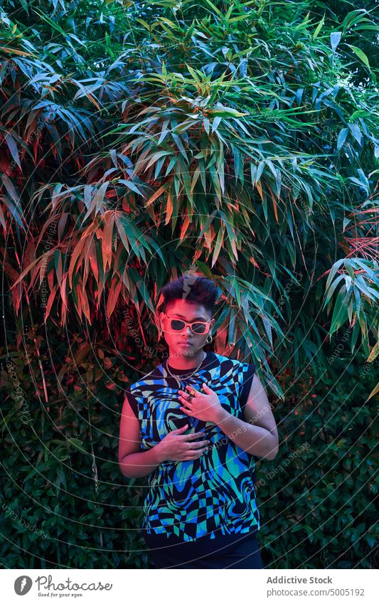 Stylish non binary person under neon light woman transgender evening bush style illuminate garden appearance sunglasses shrub feminine individuality summer