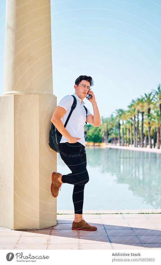 Student with backpack talking on smartphone in park student man speak call mobile tropical male gadget device using exotic pond conversation communicate