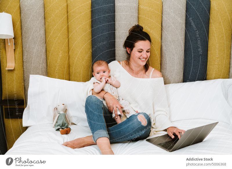 Cheerful mother using computer with interest while chilling with infant together at home laptop baby parent child cheerful bed freelance browsing surfing mom