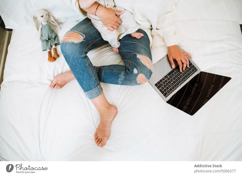 Anonymous mother using computer with interest while chilling with infant together at home laptop baby parent child cheerful bed freelance browsing surfing mom