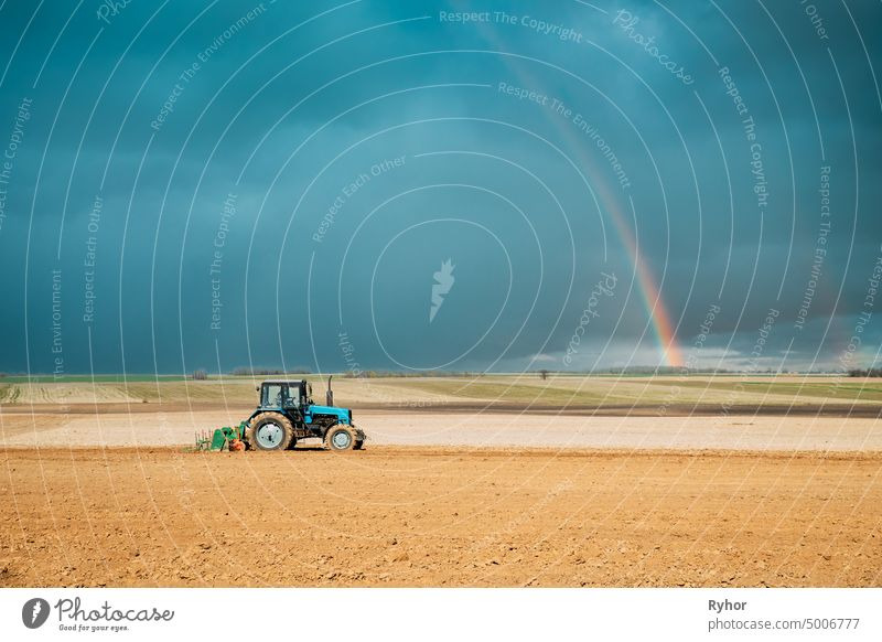 Tractor Plowing Field In Spring Season. Beginning Of Agricultural Spring Season. Cultivator Pulled By A Tractor In Countryside Rural Field Landscape Under Spring Blue Sky With Rainbow After Rain