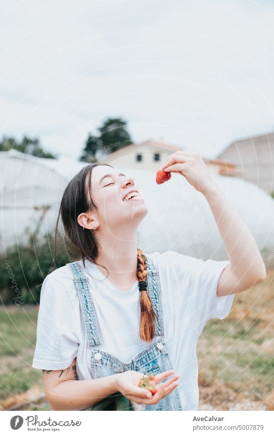 Young woman with fresh strawberries at the greenhouse, eating and playing with them, showing the fresh fruit to camera. Rural working and resources concept. hard working young woman