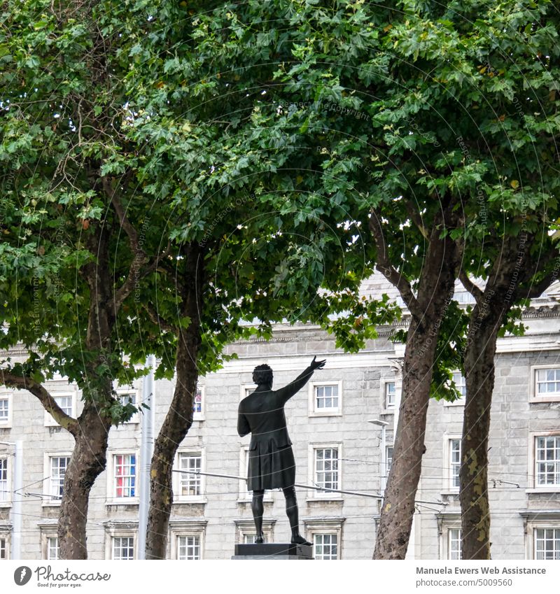 A statue under trees with raised hand in front of a big building in Dublin Statue Speaker uplifted hand Building Ireland Poet seal carry forward proclaim