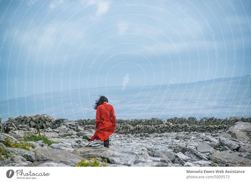 A woman with headphones walks on a stone beach, her red dress is blown away by the wind Red Dress Wind Woman stroll Going Listen to music Headphones Landscape