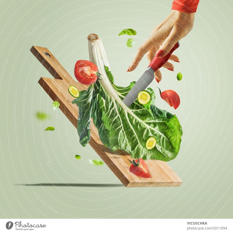 Women hand with knife , flying salad ingredients and cutting board at light green background. Creative food levitation. Healthy eating and dieting women