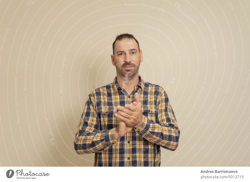 Handsome bearded latin man in a stylish checkered shirt on an isolated beige background. Clapping and clapping happy and joyful, smiling proud hands together.