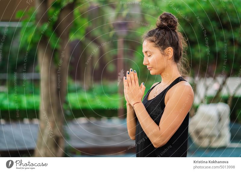 Concentrated girl sitting in lotus pose with hands in namaste and meditating or praying. Young woman with oriental appearance practicing yoga alone on wooden deck in tropical island.