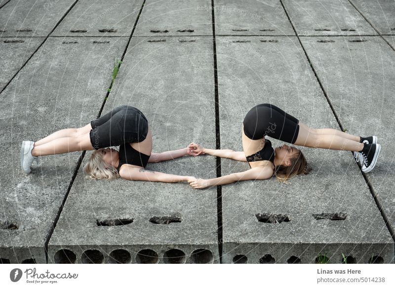 Pretty girls are holding hands while doing some kind of yoga. On a concrete. With closed eyes. These gorgeous friends are being connected and concentrated.