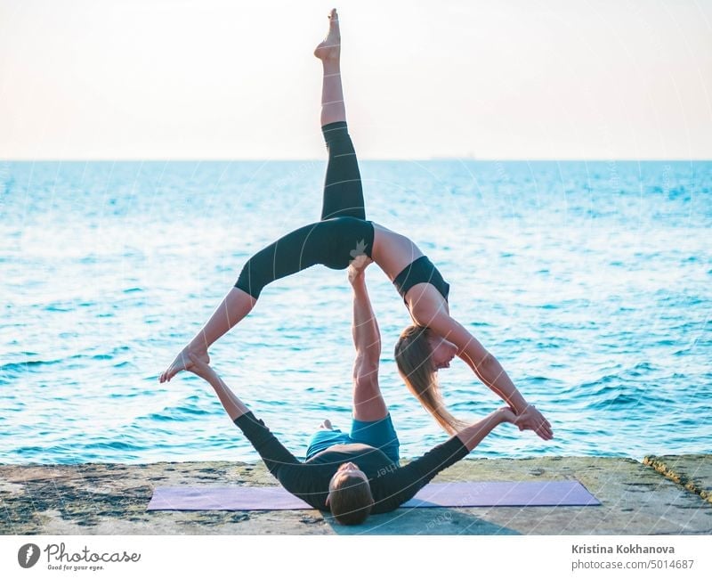 Fit young couple doing acro-yoga at sea beach. Man lying on concrete plates and balancing woman on his feet. Beautiful pair practicing yoga together. acroyoga