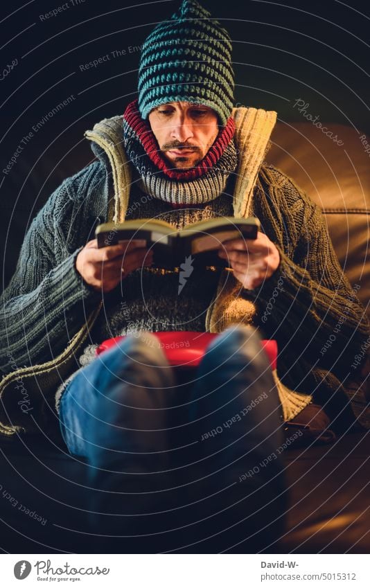 freezing at home - man wrapped in winter clothes reading a book on the sofa of his house Freeze Man inflation concept heating costs energy costs Save Cold
