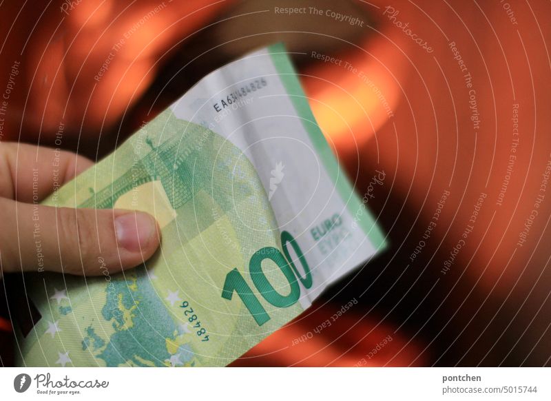 A hand holds a banknote, a hundred euros, in front of a burning fireplace. High energy company Save energy Loose change Euro finance Money Heat Fireside