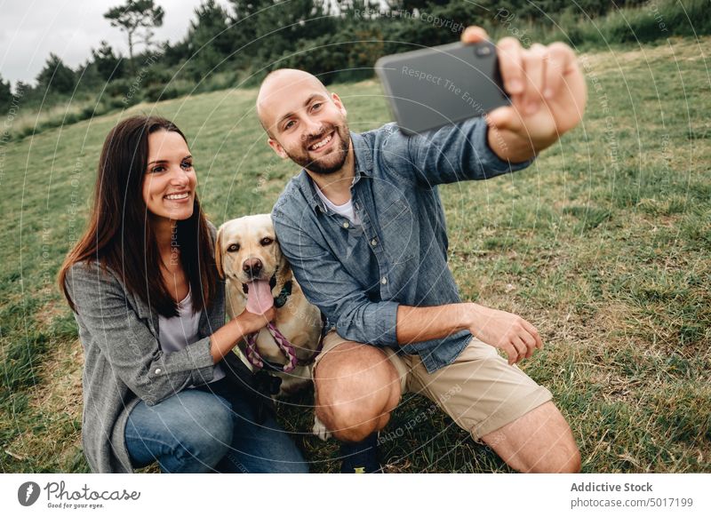 Smiling owners sitting with dog and taking selfie in park couple smartphone hugging memory using pet nature green grass animal labrador retriever joyful