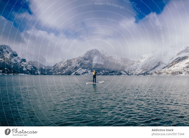 Man on paddle board between water and mountains on coast man floating tourist surface hill sup snow shore winter picturesque view stone nature travel landscape