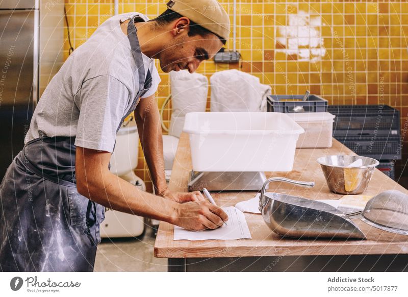Baker making notes during dough preparation bakery man work take note write prepare scale weight professional kitchen male young ethnic occupation employee