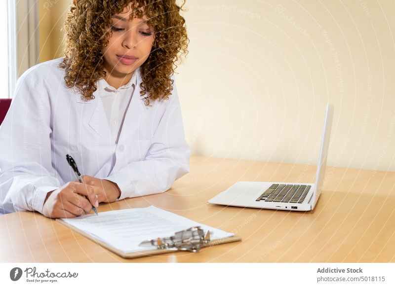 Crop ethnic medic writing on paper at table with laptop take note document professional work clipboard woman office clinic pen write doctor job attentive gadget