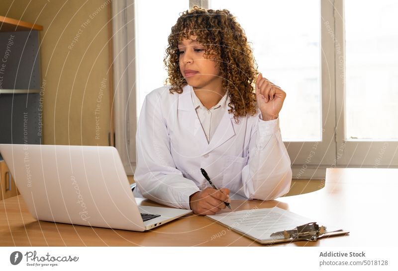 Crop ethnic medic writing on paper at table with laptop take note document professional work clipboard woman office clinic pen write doctor job attentive gadget