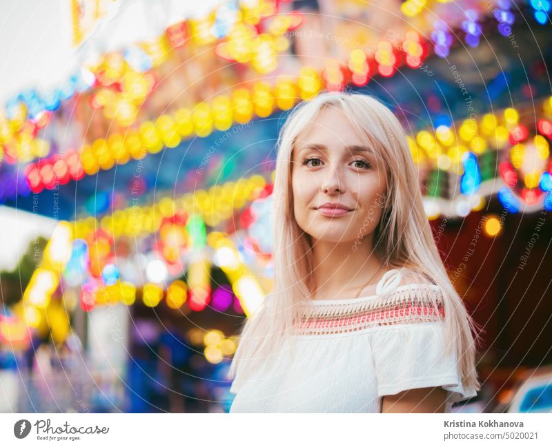 Young attractive blonde girl standing near carousel in amusement park. Beautiful portrait of woman with long hair. adult beautiful caucasian cheerful colorful
