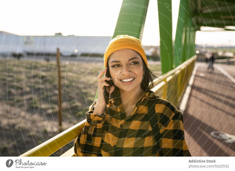Smiling young ethnic woman talking on smartphone on bridge smile style content sms positive using mobile female latin american dark hair long hair wool cap