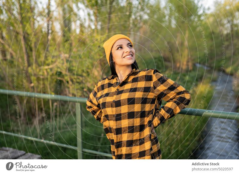 Confident young ethnic female hiker standing on wooden bridge over river in nature woman smile happy content footbridge enjoy travel forest positive