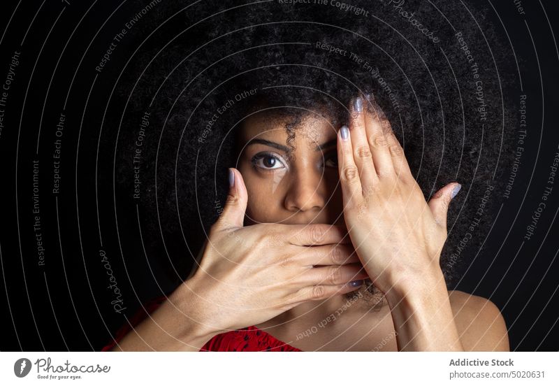 Shy ethnic woman covering face on black background cover eye cover mouth shy timid feminine hide tender afro portrait gentle hairstyle manicure romantic alone