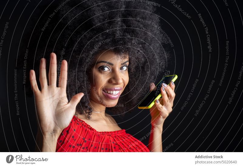 Smiling Cuban woman with smartphone saying hello on black background greeting smile feminine hand raised friendly afro portrait hi cellphone gadget black screen