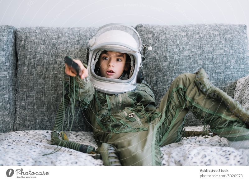 Amazed kid sitting on couch with a real astronaut uniform using a TV remote control background boy child childhood concept costume creation handmade helmet