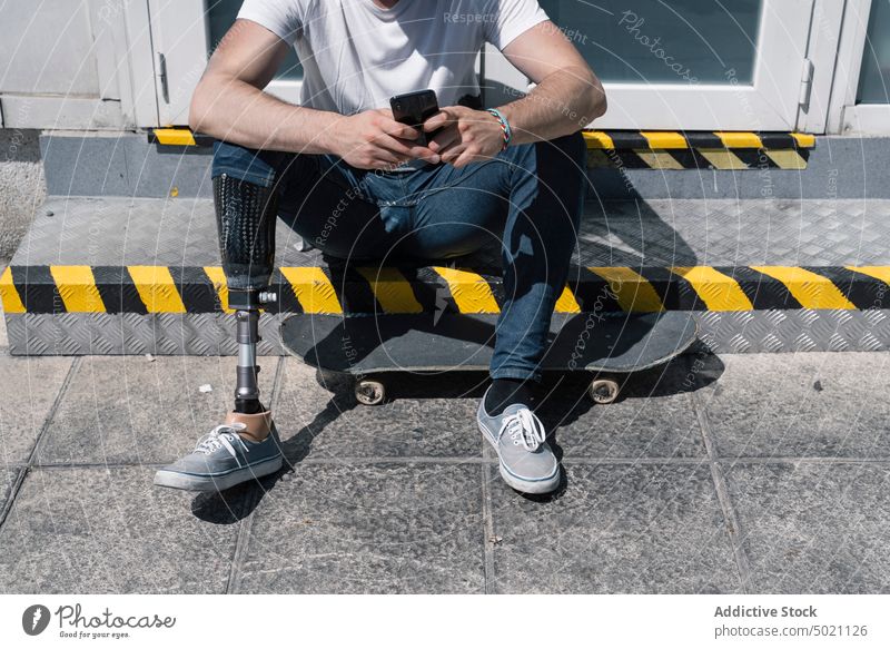 Crop skater with leg prosthesis using smartphone man skateboard artificial street city young urban male modern technology sunny daytime break social media hobby