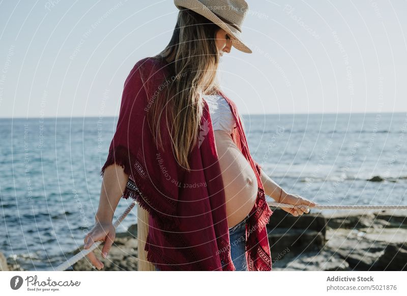 Pregnant woman resting near sea pregnant coast summer weekend vacation hat female casual blue sky beach tummy maternal daytime resort ocean shore cloudless