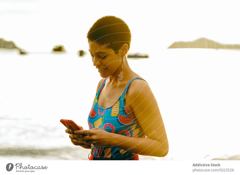 Ethnic female browsing on smartphone on beach woman summer sea communicate happy young ethnic latin american mobile smile short hair connection sunlight
