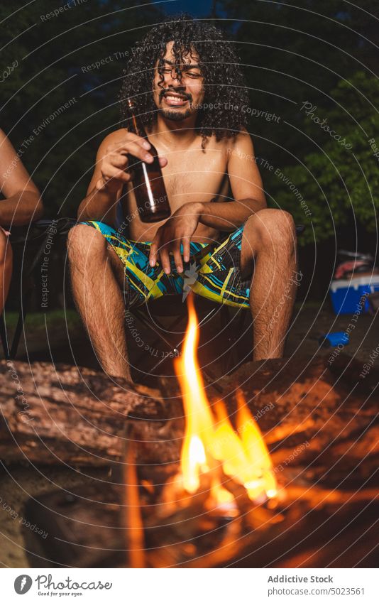 Young ethnic man drinking beer with friends near bonfire beach cheerful bottle alcohol happy evening picnic young shirtless latin american curly hair beverage