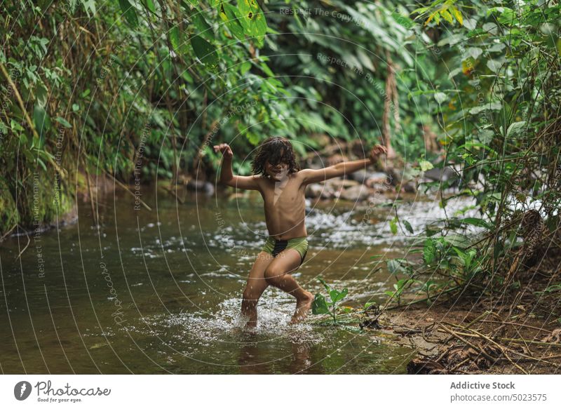 Happy ethnic boy jumping in brook in forest river joyful happy having fun kid carefree cheerful activity lush shallow woodland nature enjoy childhood energy