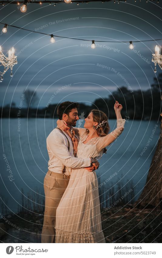 Man hugging happy woman with upped hand near tree bride groom guy lady evening wear couple chandelier field fog young embracing cheerful party smiling shindig
