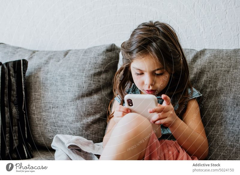 Cute ethnic child using smartphone on cozy couch at home play relax sofa comfort online internet videogame kid girl living room rest gadget casual dark hair