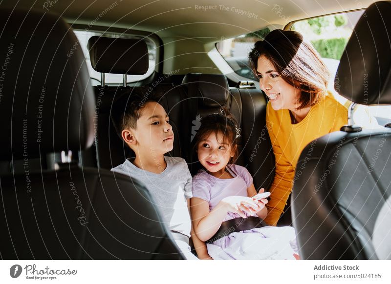Ethnic mother fastening safety belt to little girl using smartphone in car woman child daughter vehicle passenger automobile childcare motherhood childhood