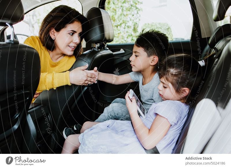 Ethnic little siblings using smartphone on car backseat and chatting with mom woman children together mother daughter son drive holding hands auto ride