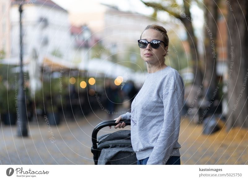 Stylish young woman wearing sunglasses walking through Ljubljana city center pushing and rocking baby stroller. Warm autumn or spring weather for outdoor activity