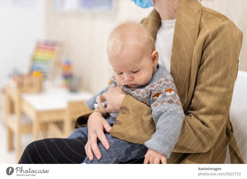 Mother holding infant baby boy in her lap, sitting and waiting in front of doctor's office for pediatric well check. child's health care concept lobby hall