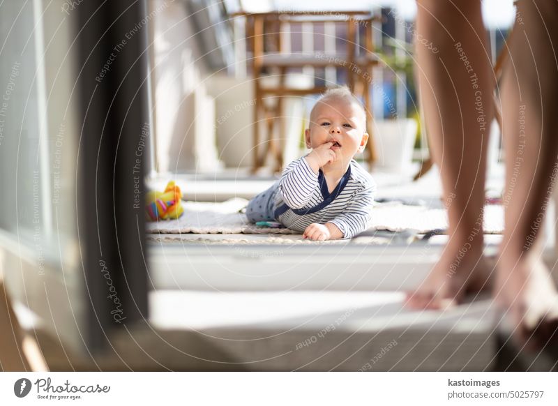Cute little infant baby boy playing with toys outdoors at the patio in summer being supervised by her mother seen in the background. childhood crawling terrace