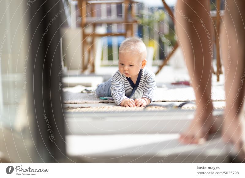 Cute little infant baby boy playing with toys outdoors at the patio in summer being supervised by her mother seen in the background. childhood crawling terrace
