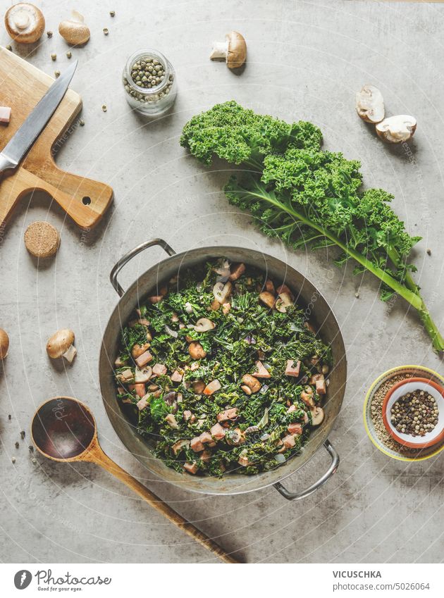 Cooking pot with stewed kale with mushrooms on kitchen table with spoon and ingredients, top view cooking pot delicious vegetable german food seasoning