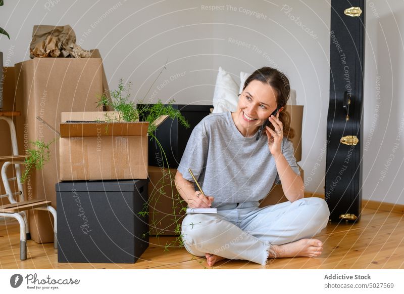 Cheerful woman speaking on smartphone during relocation write note relocate box smile homeowner happy female move in belonging rent mortgage phone call