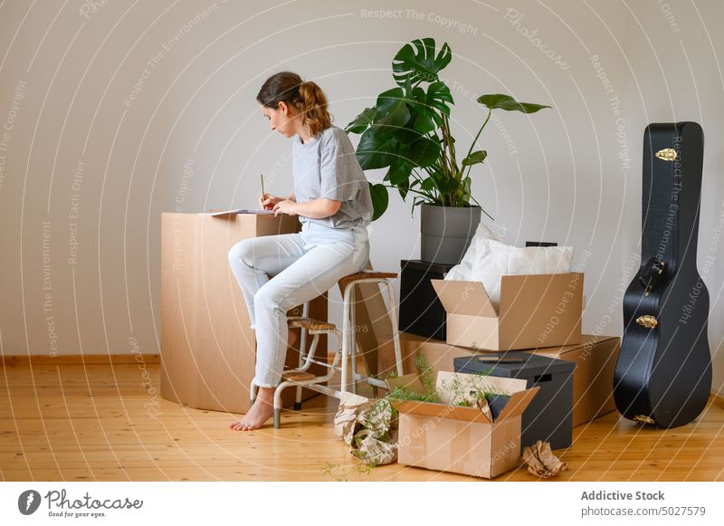 Young woman making notes on carton box write new apartment relocate homeowner ladder female move in notepad busy planner cardboard belonging occupation