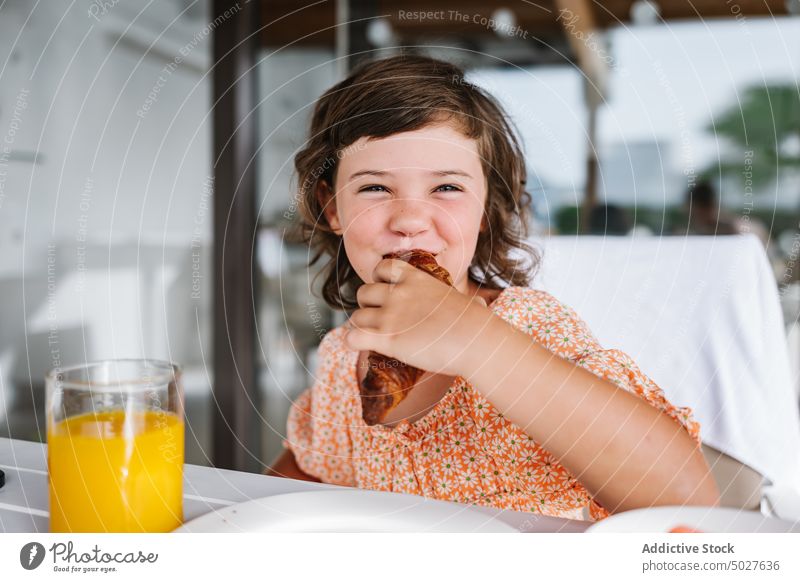 Happy kid biting tasty croissant in cafeteria girl child eat bite breakfast nutrition nourish orange juice glass food yummy natural appetizing table nutrient