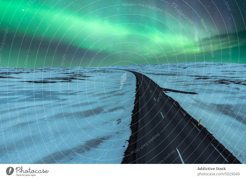 Empty asphalt road amidst field covered with snow under sky with northern lights aurora borealis winter landscape night polar Iceland breathtaking ridge