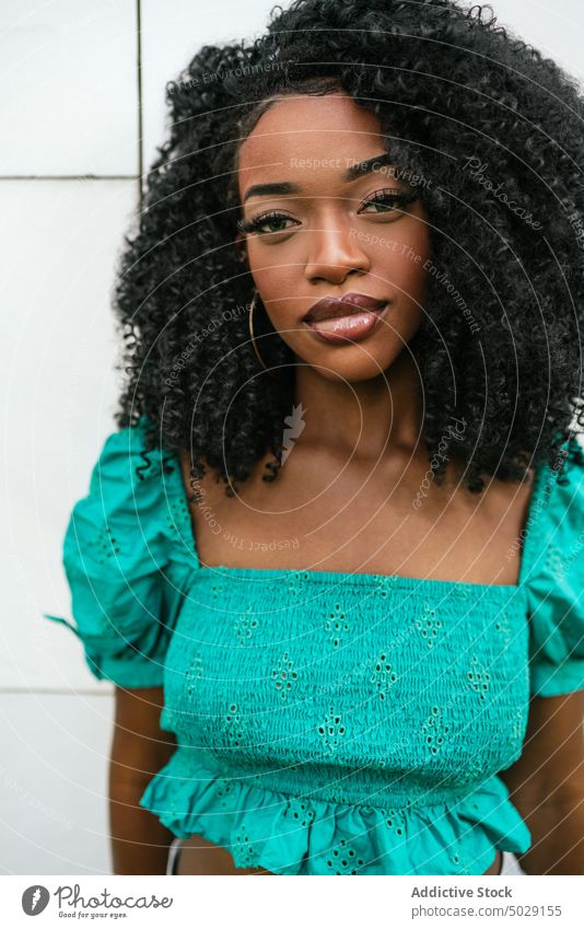 Serious African American woman standing near white wall unemotional model style appearance top turquoise afro makeup casual hairstyle outfit confident tile slim