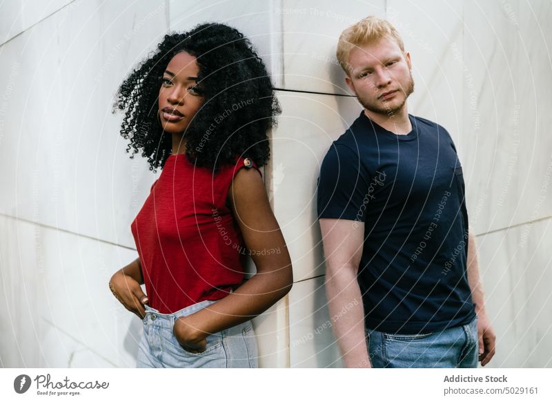 Serious multiethnic couple standing near white building in park boyfriend girlfriend serious unemotional lean on tile together appearance hairstyle casual afro