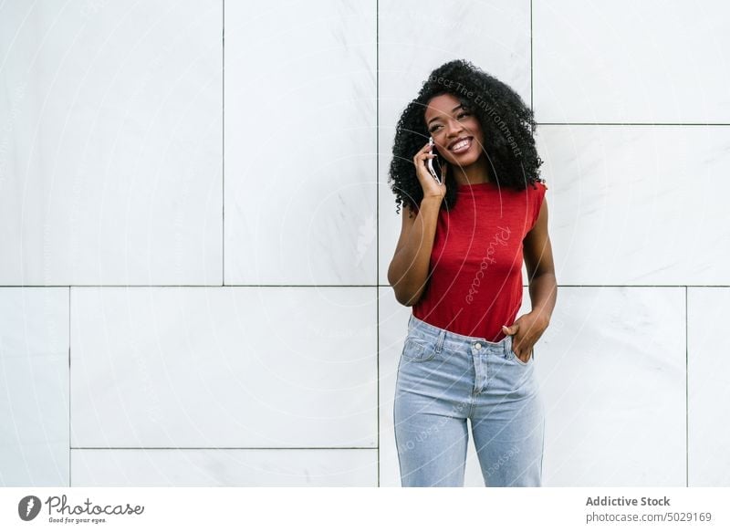 Cheerful African American woman talking on smartphone near wall cheerful smile cellphone hand in pocket speak phone call street happy style device conversation