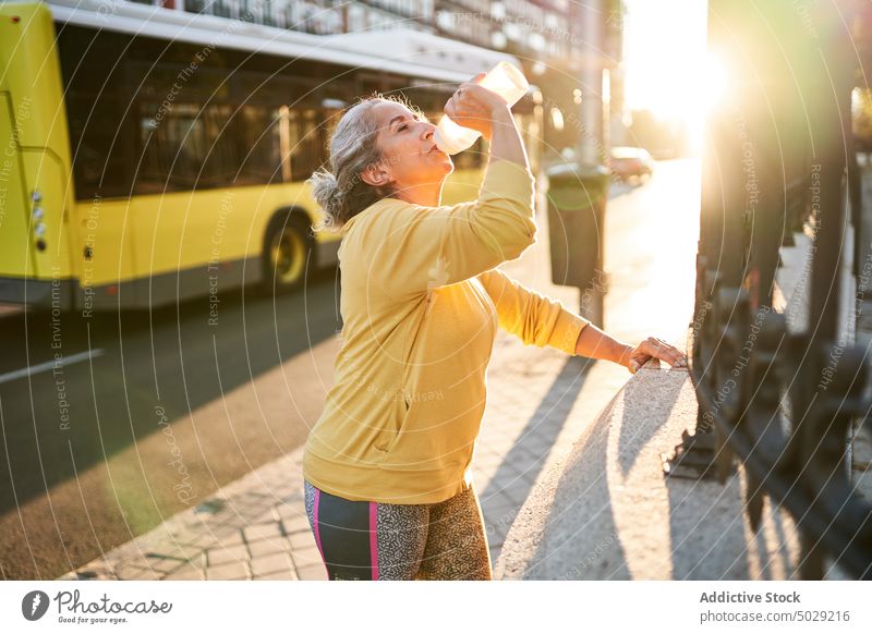 Aged sportswoman taking break on street drink fitness training sunrise bottle water fence female elderly senior aged wellness energy power pensioner wellbeing