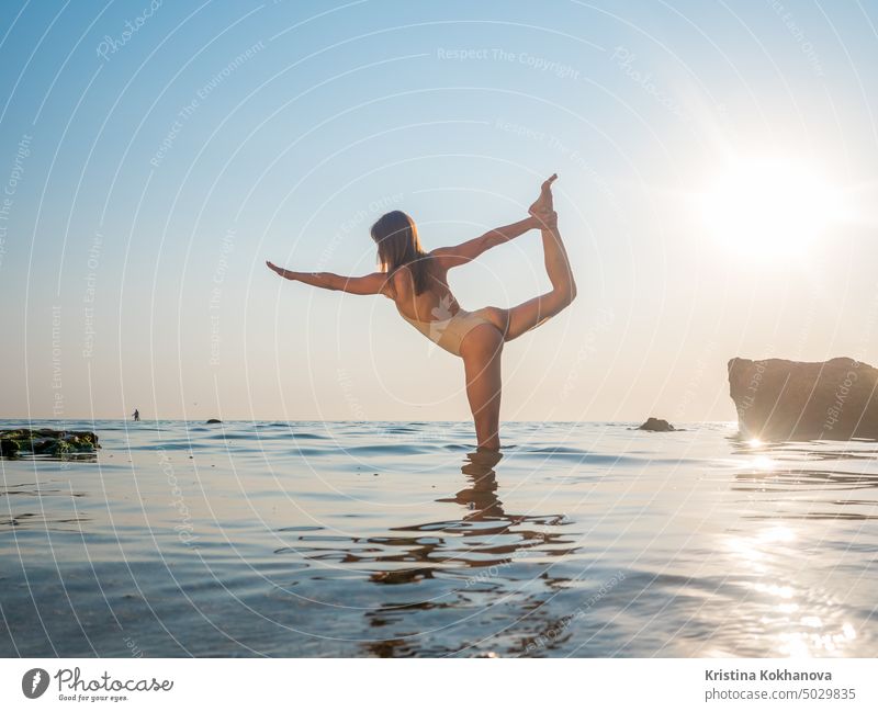 young woman in swimsuit practicing yoga at amazing sunrise. Fitness, sport, yoga and healthy lifestyle concept. Girl doing exersices in sea water nature sunset