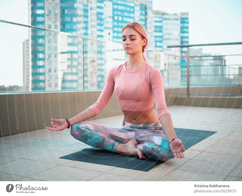 Young beautiful woman in pink tracksuit meditating during outdoor training on skyscraper terrace, she feeling peace in megalopolis. Sporty girl doing yoga on the roof.